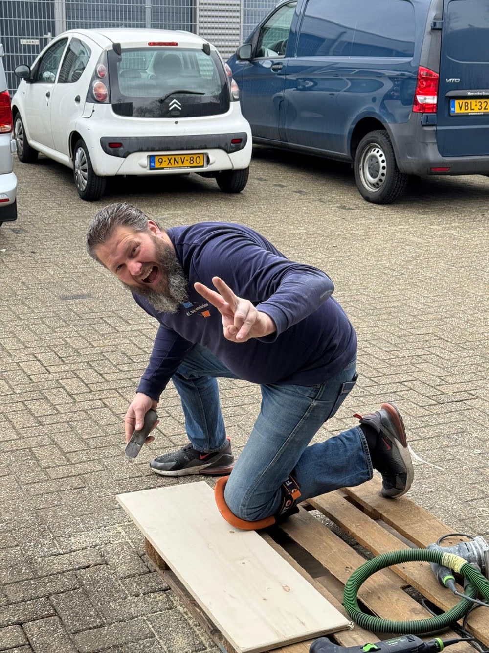 Herman op de parkeerplaats bij de werkplaats, met een grijns en een vredesteken